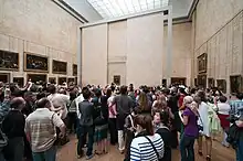 Farbfotografie von einer großen Besuchermenge, die Mona Lisa im Hintergrund fotografiert. Das Gemälde ist hinter Glas in einer Mauerkonstruktion gehängt, die frei im Raum steht. Weitere alte Gemälde befinden sich an den Wandseiten des Raums, der von einem breiten Deckenlicht beleuchtet wird.