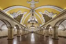 Frontale Farbfotografie einer U-Bahn-Station mit gelbem Gewölbe und weißen Ornamenten, die über einen langen Gang bis zum Hintergrund führen. An beiden Seiten des Gangs sind Pfeiler und Gleise. In der Mitte der Decke hängen Kronleuchter und an den Seiten der Decke sind goldbraune Wandmalereien.