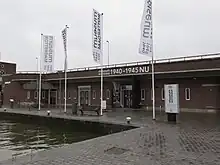  Eingangsbereich des Museum Rotterdam ’40 – ’50 NOW am Hafenbecken, mit mehreren Fahnen und einer Sitzbank vor dem Eingang