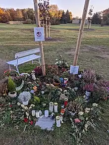 Viele Blumen und Kerzen. Auf einer großen Wiese, im Hintergrund andere Bäume. Am Ort fehlt der Baum.