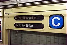 Detailansicht im Inneren eines U-Bahn-Wagens: Ein begie lackierter Metallkasten mit drei verglasten Öffnungen zeigt in gedrängter Form Informationen zum Zuglauf an. Links oben steht „168 St, Manhattan“, links unten steht „Euclid Av, Bklyn“, beides in weißer Schrift auf schwarzem Rollband. Rechts ist groß das runde blaue Linienemblem mit einem weißen Großbuchstaben C zu sehen.