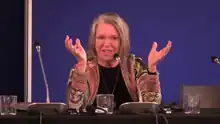 Nancy Fraser, mit grauen Haaren und Brille sitzt hinter einem Mikrofon auf einem Podium.