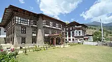 Ansicht des Gebäudes der Nationalbibliothek von Bhutan. Ein Gebäude mit einer hohen zweigeschossigen Wand aus gemauertem Naturstein und zwei Obergeschossen aus Fachwerk.