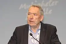 Foto eines älteren, anzugtragenden Mannes, der in ein Mikrofon spricht