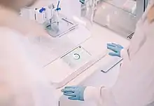 Eine Person im weißen Kittel und mit blauen Gummihandschuhen steht vor einem Labortisch, auf dem ein 3D-Bioprinter steht. Dieser besteht aus einer vertikal ausgerichtet Spritze, Linearantrieben und einer Petrischale