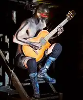  Riedel sitzt auf einer Bühnentreppe mit akustischer Gitarre. Der Oberkörper ist unbekleidet, der rechte Arm ist mit schwarzem Klebeband beklebt und der Kopf schwarz geschminkt mit einem roten Augenstreifen.