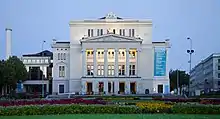 Frontale Farbfotografie von einem weißen Gebäude mit einem breiten Mittelteil, das noch ein zusätzliches Geschoss hat. Das Mittelgeschoss besteht aus Säulen und einem Giebelfeld mit einer Szene. Unter dem Giebelfeld steht „Nacionale Opera“. Am rechten Seitenteil hängt ein langes Banner mit dem Programm vom August. Auf dem Vorplatz sind Blumenbeete.