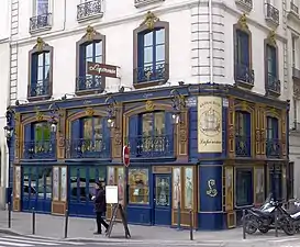 Seitliche Farbfotografie eines Gebäudes, von dem nur die ersten drei Stockwerke zu sehen sind. Erdgeschoss und erstes Geschoss haben eine dunkelblaue Fassade mit gemalten Menschen aus dem 19. Jahrhundert und braun-goldenen Ornamenten zwischen den Fenstern. An der Ecke steht um ein gezeichnetes Segelschiff herum „Restaurant Lapérouse“ und der Name „Lapérouse“ ist auf einem Aushängeschild im zweiten Geschoss, dessen Fenster braun-gold umrandet sind.