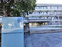 Ein dreistöckiges, graues Gebäude aus Beton mit dem Namen "Pädagogische Hochschule Vorarlberg" buchstabiert in silbernen Buchstaben an der Fassade und einem Schild mit der PH Vorarlberg-Logo im Vordergrund.