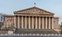 Frontale Farbfotografie eines klassizistischen Gebäudes mit einem breiten Säuleneingang und fünf grünen Glastüren. Über den Säulen steht in Gold „Assemblée Nationale“ und im Giebelfeld ist eine antike Szene um einen Thron. Auf dem Dach ist eine Frankreichflagge und links neben dem Gebäude wehen drei Frankreichflaggen und eine Europaflagge. An beiden Seitenmauern sind in schmalen Rechtecken antike Szenen. Links und rechts neben den Außenstufen sind Steinfiguren.