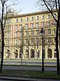 Frontale Farbfotografie einer Gebäudefassade mit fünf Stockwerken. Im Erdgeschoss bestehen die Türen und Fenster aus Rundbögen. Der Mittelteil des ersten und zweiten Stockwerks ist mit Säulen verziert und auf einem Balkonende stehen zwei Figuren. Zwischen den Fenstern im Obergeschoss befinden sich Ornamente und auf der linken Dachseite ist eine Kuppel. Der Vordergrund ist von einer Straße und kahlen Bäumen durchzogen.