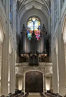 Das Foto zeigt den Blick aus einiger Entfernung auf die Hauptorgel und die zugehörige kleine Brüstungsorgel auf der Empore der Kirche Saint-Gervais. Hinter der Orgel sieht man Teile eines hohen bunten Kirchenfensters, das in den Farben Gelb, Grün, Rot, Blau und Weiß im Gegenlicht leuchtet; im Vordergrund des Fotos sieht man hohe weiße Pfeiler und einige Reihen mit dunklen hölzernen Sitzhockern.
