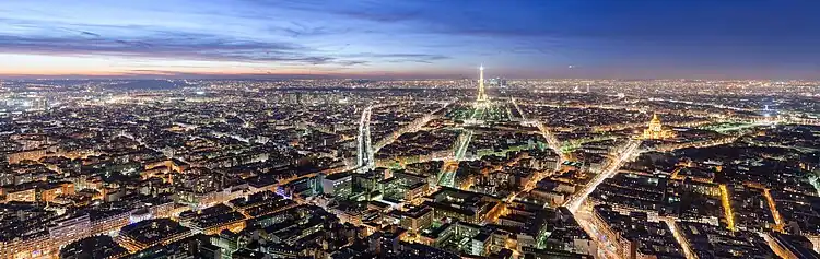Nachtaufnahme vom beleuchteten Paris mit dem Eiffelturm und dem Pantheon auf der rechten Seite. Der blau-violette Himmel hat links die gelben Reststreifen der untergehenden Sonne und rechts einen weißen Lichtstreifen.