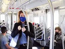 Feinberg zwischen Reihen von Haltestangen stehend in einem U-Bahn-Zug mit mehreren sitzenden Fahrgästen bei Tageslicht, Stoffbeutel mit Gesundheitsartikeln für die Fahrgäste über der Schulter, Mobiltelefon in der Hand