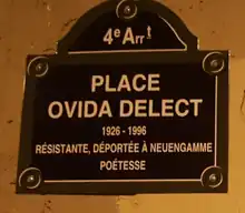 Auf dem Foto ist an einer sandsteinfarbenen Wand eine dunkelbraune Tafel mit goldfarbenen Buchstaben befestigt. Dort steht auf französisch: Viertes Arrondissement. Place Ovida Delect (1926–1996). Widerstandskämpferin, nach Neuengamme Deportierte, Dichterin.