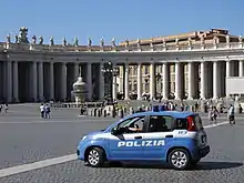 Farbfotografie von einem riesigen Platz, der von einem hellen Säulengang mit Figuren drauf umschlossen ist. Verteilt laufen Menschen auf dem Platz, das einen Springbrunnen hat. Im Vordergrund ist ein blau-weißer Polizeiwagen mit der Aufschrift „Polizia 113“ zu sehen. Im Wagen sitzt ein Mann, dessen Arm im Licht gut zu erkennen ist.