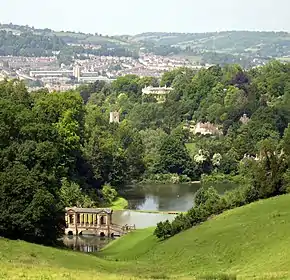 Farbfotografie in der Obersicht eines Parks mit vielen Bäumen, zwischen denen alte Gebäude und Türme stehen. Im Vordergrund ist eine hügelige Wiese, die sich an einen Weiher mit Bäumen anschließt. Über die vordere Wasserfläche führt eine antik aussehende Brücke mit Überdachung. Im linken Hintergrund ist ein Ort, der von einer Hügellandschaft aus Bäumen und Wiesen umgeben ist.