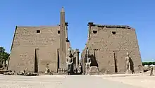 Paar trapezförmiger Steintürme, die einen Durchgang flankieren, hinter dem eine Säulenreihe sichtbar ist. Vor den Türmen stehen mehrere große Statuen und ein Obelisk.