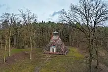 Eine vierseitige Natursteinpyramide mit einer überdachten Spitze und einem hellen Eingangstor steht auf einer Waldlichtung, umgeben von Mischwald.