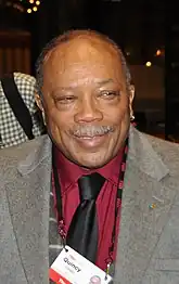 An older man with a white mustache, wearing a black tie, red dress shirt, gray jacket, and a name badge that hangs from a string around his neck.