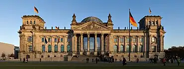 Frontale Farbfotografie vom Reichstagsgebäude bei Sonnenuntergang. Ein breiter Treppenaufgang führt zum Glaseingang mit Steinsäulen im Vordergrund. Über dem Eingang steht unter einem Giebelfeld mit Szene in alter Schrift „Dem deutschen Volke“. Mittig auf dem Gebäude ist die Glaskuppel. Auf beiden Seiten des Daches sind zusätzlich rechteckige Geschosse mit je einer Deutschlandflagge.