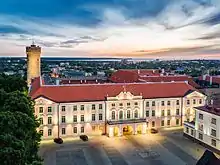 Farbfotografie bei Nacht von einem hellen Gebäude mit weißen Verzierungen. Das dreigeschossige Gebäude hat in der Mitte drei Torbögen als Eingang und ein Giebelfeld mit goldenem Wappen. Zwei Flaggen wehen auf dem braunen Schrägdach. Im linken Hintergrund steht ein Turm.