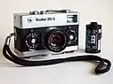 Frontale Farbfotografie einer analogen Kamera der Marke Rollei 35 S mit einer Schlaufe, die im Vordergrund liegt. Die Kamera hat neben dem Objektiv zwei Drehräder und  auf der Kamera sind zwei Knöpfe und ein Spannhebel. Rechts steht eine Filmrolle.