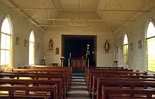 Das Foto eines Innenraums einer Holzkirche. Der Betrachter steht in der Kirche und blickt zwischen den beiden Bankreihen entlang auf den Holzaltar. Dieser steht in einer Nische. Rechts und links von der Nische befindet sich je eine Heiligenfigur an der Wand. Vor dem Altar steht ein Rednerpult. Die Wände und das Dach der Kirche sind weiß gestrichen und spärlich mit Bildern, deren Inhalt nicht erkennbar ist, dekoriert. Durch die schlichten Glasfenster auf der linken Seite scheint die Sonne in den Innenraum.