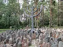 Frontale Farbfotografie von einem Feld aus mittelgroßen Steinen, die um ein metallenes Kunstwerk stehen. Das Kunstwerk besteht aus sich schlängelnden Drähten und soll den Chanukkaleuchter darstellen. Die Drähte verbinden sich mit den Steinen und symbolisieren Baumwurzeln. Im Hintergrund ist Wald.