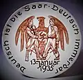 Gedenkteller zur Saarabstimmung: Brauner Reichsadler mit Hakenkreuzemblem, davor reichen sich ein NS-Parteigenosse (links) und ein Saarbergmann mit Grubenlampe (rechts) die Hand. Unterhalb ist das Datum der Abstimmung eingetragen: „13. Januar 1935“: Entlang des Randes befindet sich die Umschrift „Deutsch ist die Saar – Deutsch immerdar“ nach dem „Saarlied“ von Hanns Maria Lux. Waechtersbacher Steingut, 1935