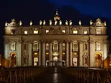 Frontale Nachtfotografie von einem Renaissance-Gebäude mit Säulen, deren Fenstern leuchten. Die Figuren auf dem Dach und die Turmspitze im Hintergrund sind angestrahlt. Im Vordergrund sind an den Seiten Holzabsperrungen.