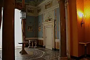 Farbige Eckansicht eines Innenraums mit zwei Säulen im vorderen Durchgang. Die hellblauen Wände sind mit goldenen Ornamenten und kleinen Gemälden bemalt. Über der weiß-gold verzierten Tür an der rechten Wand ist ein helles Rundbogengemälde gezeichnet. Die oberen Wandseiten sind mit hellen Ornamenten und Gemälden mit seitlichen Holzläden gemalt. Links hinten ist ein Fenster mit braunen Gardinen zu sehen. Im Raum stehen zwei Beistelltische und rechts strahlt eine Wandleuchte.