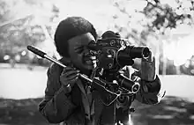 Schwarz-Weiß Fotografie einer Schwarzen Frau mit kurzen Haaren, die eine altmodische Filmkamera führt. Ein Auge ist am Sucher der Kamera, das andere ist zugekniffen.