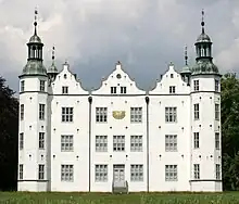 Frontale Farbfotografie eines dreigeschossigen Schlosses mit einer Sonnenuhr im Obergeschoss und vier Ecktürmen, auf denen Turmhauben sind. Zwischen den vorderen Türmen befinden sich drei Staffelgiebel. Im Vordergrund steht eine Wiese und an beiden Seiten sind Bäume zu sehen.