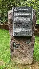 Auf der Steintafel eingravierter Text: „Schnullerbaum. Ein Schnullerbaum dient der einfacheren Schnuller-Entwöhnung eines Kleinkinds. Die Idee des Schnullerbaums stammt aus Dänemark. Auf ausgewiesenen Bäumen können Kleinkinder ihren Schnuller aufhängen, um sich so leichter von diesem zu lösen. Der Gedanke ist, dass das Kind die sonst eher problematische Trennung vom Schnuller mit einem positiven Erlebnis verbindet, da es den Schnullerbaum jederzeit besuchen und auf diese Weise auch an die Natur herangeführt werden kann. Es sieht, dass auch andere Kinder ihren Schnuller abgegeben haben. Im Anschluss erhält es meist ein kleines Geschenk oder eine Urkunde. Verschönerungsverein Gönnern“