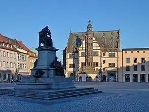 Im Vordergrund eine im Schatten liegende auf einem steinernen Podest stehende Statue. Am Podest sind zwei weitere Statuen platziert. Dahinter steht neben anderen, von der Sonne beschienen Häusern zentral ein mit Wappen verziertes fachwerkartiges Gebäude mit einem zentralen Turm und einer Uhr.