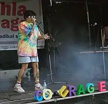 Schwesta Ebra steht auf einer Bühne und trägt ein buntes Batik-Shirt und eine kurze helle Hose. Dazu trägt sie weiße Nike-Sneaker und Socken, mit Regenbogenfarben. Sie hat kurzes schwarzes Haar und trägt eine weiße Sonnenbrille. Im Vordergrund sind bunte Buchstaben zu sehen, die das Wort Courage bilden.
