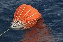 Ein oranger falschirmähnliches Tuch mit Leinen daran liegt im Wasser und wird offenbar hinter einem Schiff nachgeschleppt