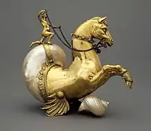 Vergoldete Figur, deren vordere Hälfte wie ein Pferd aussieht, an dessen Bauch eine kleine Flosse ansetzt, die hintere Hälfte aber aus einem perlmuttfarbenen Meeresschneckenhaus besteht, auf dem eine kleine menschliche Gestalt kniet und Zügel in den Händen hält.