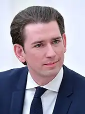 Porträtfoto, darunter Unterschrift. weiße glatte Haut (rasiert), Kopf gedreht und schräg, blauer Anzug, weißes Hemd, blaue Krawatte mit kleinen weißen Punkten, nach hinten gekämmtes schwarz-graues Haar, lächelt kaum, insgesamt ordentlicher Eindruck.