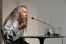 Seitenansicht eines Mannes mit langen grauen Haaren, er sitzt an einem Tisch mit Wasserkaraffe und spricht in ein Mikrofon.