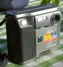 Farbfotografie einer Digitalkamera mit der Aufschrift Sony Mavika. Das linke Gehäuse ist schwarz, der Rest hat die Farbe Silber. Links ist ein Knopf und rechts ist das Objektiv.