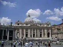 Frontale Farbfotografie vom Petersdom mit dem Platz und vielen Menschen im Vordergrund. Ein Teil des linken Säulengangs vom Platz ist zu sehen. Die Säulen verlaufen über zwei Geschosse. Auf dem Dach sind Marmorfiguren und eine Kuppel.