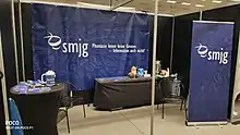 Zu sehen sind Banner mit der Aufschrift „smjg: Phantasie kennt keine Grenzen – Information auch nicht!“, ein Tisch mit Printmedien zum Mitnehmen und ein Tisch mit Kaffeemaschine, Bechern und Stofftier-Maskottchen