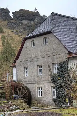 Blick auf den Nordgiebel der Stoffelsmühle. Davor das 2011 installierte Wasserrad; oberhalb der Mühle die Radfahrerstatue von Kleinziegenfeld