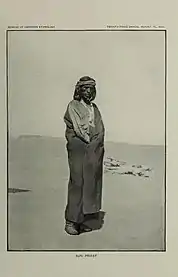Schwarz-Weiß-Fotografie eines Sonnen-Priesters der Zuñi, der in einer kargen Wüstengegend steht. Er trägt ein graues Band um die Stirn, ein bis zu den Füßen reichendes, dunkles Gewand, ein nur teilweise sichtbares helles Hemd, eine weiße Halskette, an der zwei spitze Steine hängen, und dunkle Arbeitsschuhe. Seine schwarzen Haare trägt er als die Ohren verdeckende Bobfrisur.