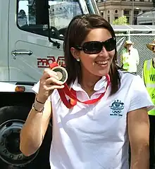 Suzie Fraser trägt ein weißes T-Shirt und eine Sonnenbrille. Die Medaille ist an einem roten Band befestigt. Im Bildhintergrund ist ein Truck zu sehen, daneben stehen zwei Ordner.