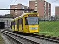 Gelber Straßenbahntriebwagen fährt auf zweigleisiger Strecke von links aus einem betonierten Tunnelportal eine leichte Rampe in Richtung des Fotostandorts hinauf. Im Bildhintergrund backsteinfarben verschalte mehrgeschossige Wohnblöcke im Stil der 1970er Jahre.