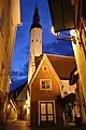 Nachtaufnahme in der Untersicht von einer Kirche in einer kleinen Gasse. Der schmale, weiße Kirchturm mit schwarzer Turmlaterne ragt bis zum oberen Bildrand. An der Kirche steht ein rotes Vorderhaus mit Wappen über der Tür. Die Gasse führt links an der Kirche vorbei und die Häuser sind mit strahlenden Laternen ausgestattet.