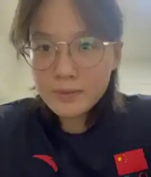 Tang Muhan trägt einen schwarzen Trainingsanzug mit der roten Fahne der Volksrepublik China und eine Brille.